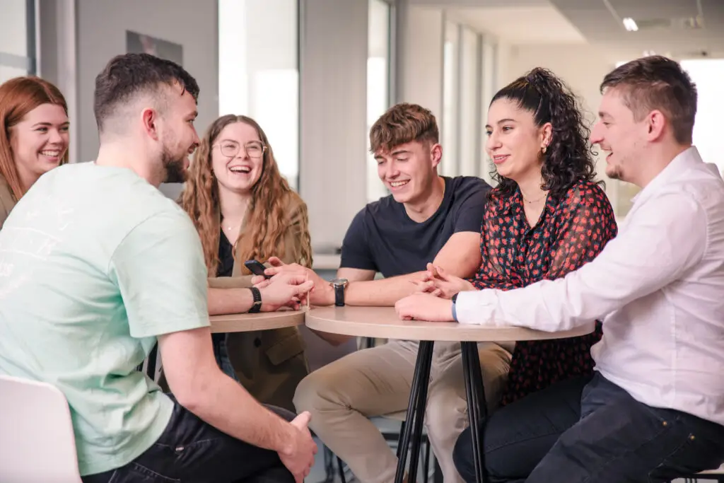 VIGNETTE – MAS MANA STRAT Photo de six étudiants autour d'une table haute dans une salle de pause, rigolant tous ensemble.