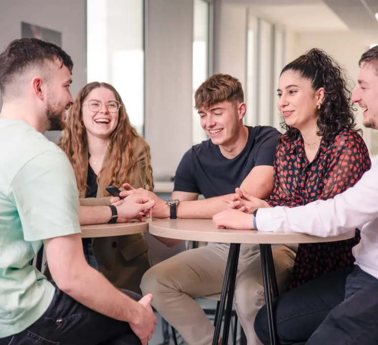 VIGNETTE – MAS MANA STRAT Photo de six étudiants autour d'une table haute dans une salle de pause, rigolant tous ensemble.