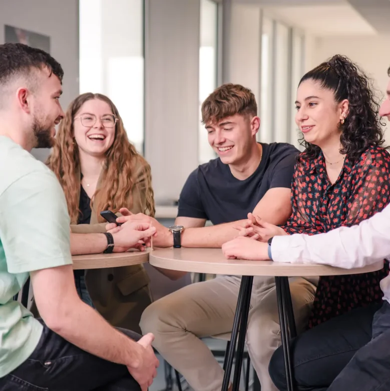 Photo de six étudiants autour d'une table haute dans une salle de pause, rigolant tous ensemble.